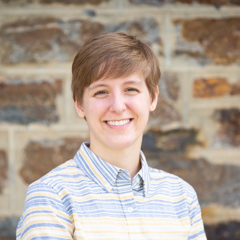 A headshot of Kirsten. She has light skin, short blond hair, and is smiling. Kirsten is wearing a white, yellow, and blue striped button-up shirt and is in front of a brick wall.