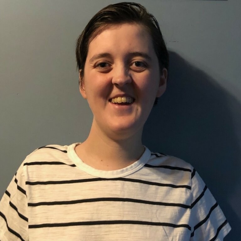 A portrait of Emily who has pale skin, short brown hair, and brown eyes. She is smiling and wearing a cream-colored shirt with navy blue stripes. The wall behind her is a light blue.