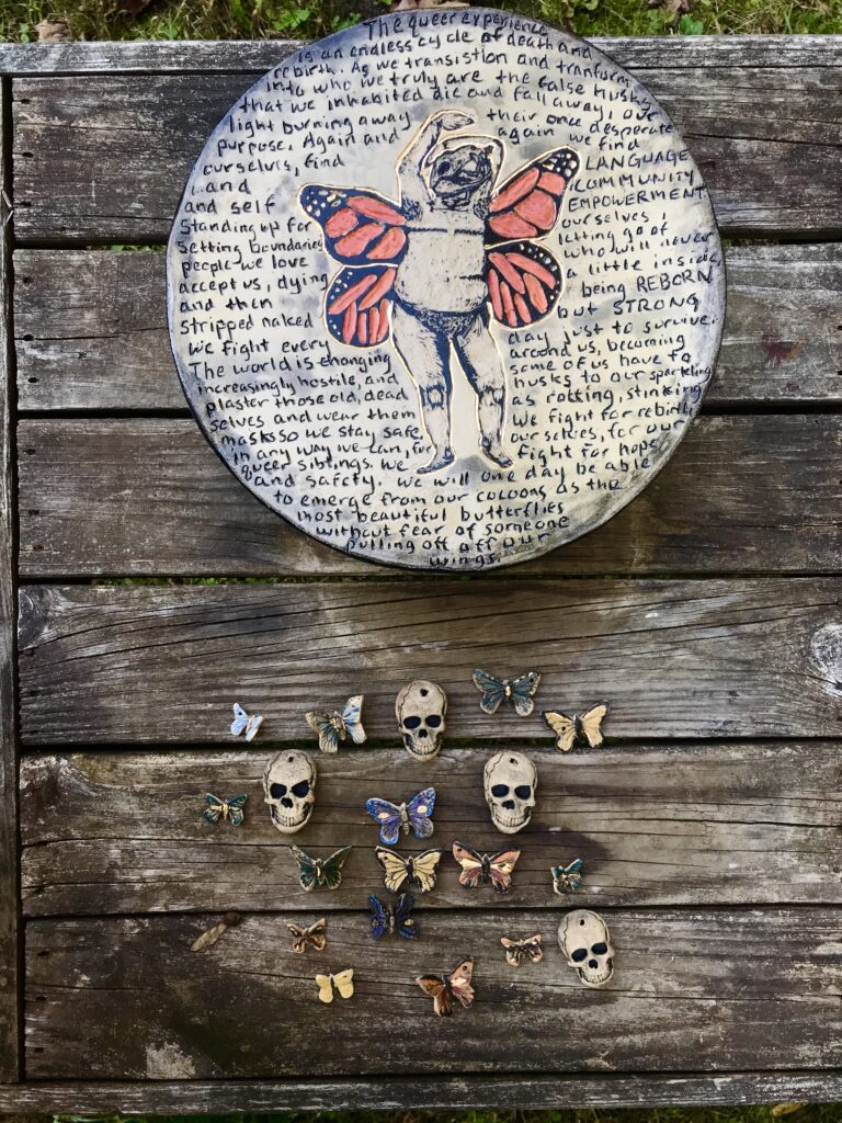 A round ceramic artwork with a person with butterfly wings and a poem surrounding them. Small skulls and butterflies hang from the bottom of the piece. 