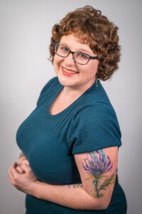 A photograph of Ali Kane smiling at the camera with hands clasp at her waist against a gray background. Ali has pale skin, cropped, curly auburn hair, and blue plastic frame glasses. She is wearing a blue short-sleeved sweater and has a large watercolor thistle tattoo on her upper arm.
