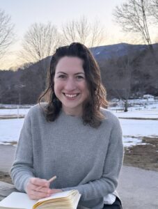 Patty has curly shoulder-length brown hair, pale skin, and is smiling. Patty is wearing a gray sweater and is sitting outside at sunset with mountains in the background and a sketchbook in her hand. 
