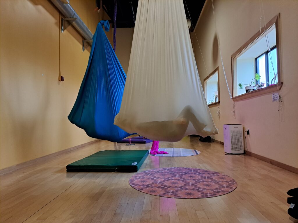 A yoga studio. Hanging from the ceiling are large hammock structures. There are outlines of bodies resting in the hammocks.