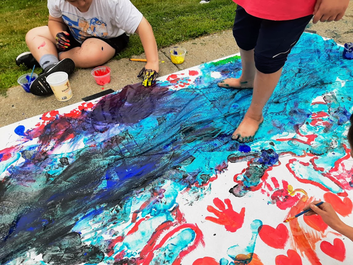 Two children are outdoors painting with their fingers and toes. One is standing on a large canvas with feet covered in red and blue paint and the other is seated on the ground with legs crossed and hands covered in paint, with one hand placed on the canvas. The canvas itself is coated in many streaks, swirls and waves of blue and red paint, which in some spots has blended to create a purple hue. For privacy purposes, the children’s faces are not visible, but their joy for the artmaking process shines through their engagement in the creative process.