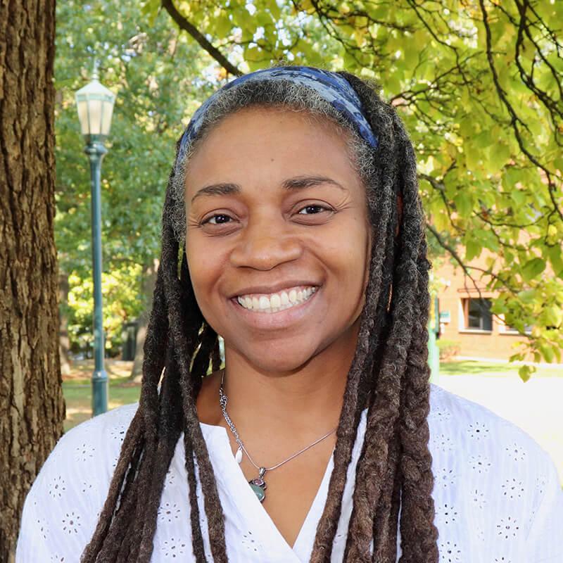 A portrait of Dr. Looby, a Black woman with long hair in locs and a blue headband on top of her head. She is smiling and wearing a white shirt with small embroidered flowers and a silver necklace. Behind her are trees with green leaves. 