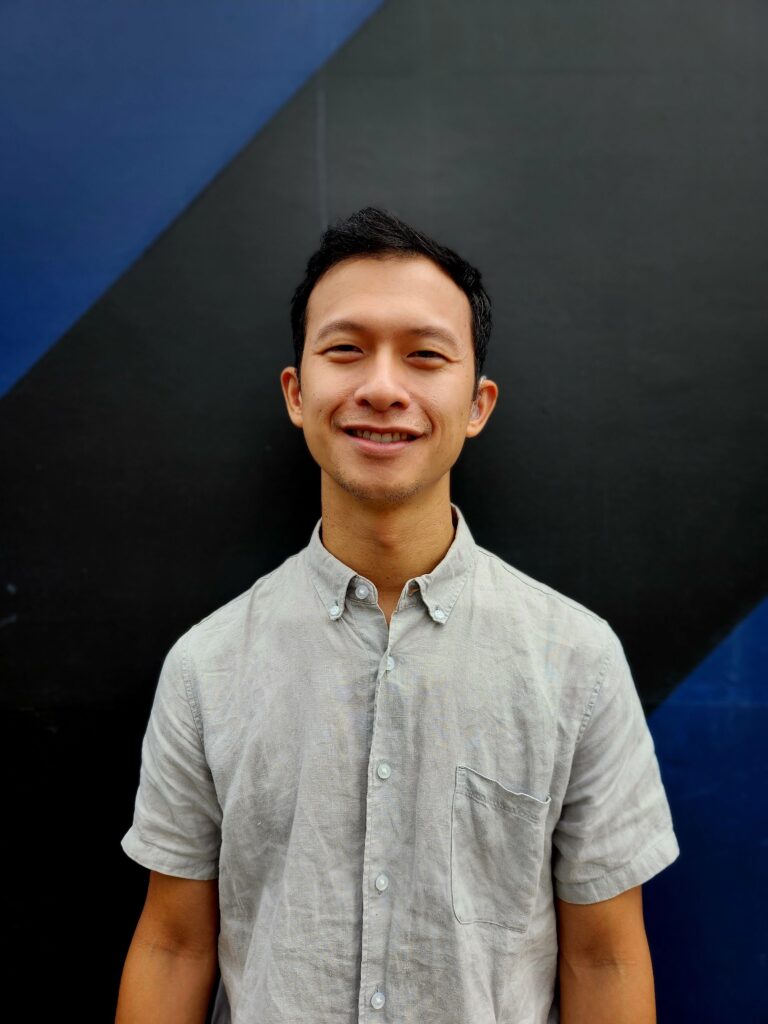 A portrait of John, an East Asian-American man with short black hair. He is smiling and wearing a grey linen button-up shirt. On his left ear, there is a clear-colored hearing aid. Behind him is a wall painted with large diagonal blue and black stripes.