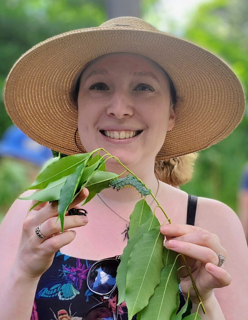 A portrait of Judith, a white woman with pale skin and blue eyes. She is smiling and wearing a wide-brimmed straw hat and a black tank top with a jewel-tone insects design, including moths and beetles. She is holding a plant stem with lots of green leaves and a large green caterpillar crawling across the top.